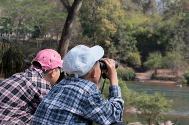 Asian boys are using binoculars to do the birdwatching in national park during their summer vacation, idea for learning creatures, wildlife animals, insects, trekking and hiking outside the classroom.