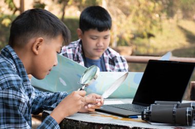Asian boys hold bird feather and binoculars sit together in local national park to learn differentces of birds from feather during their summer vacation, leisure and outdoor activity of teens concept.