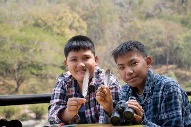 Asian boys hold bird feather and binoculars sit together in local national park to learn differentces of birds from feather during their summer vacation, leisure and outdoor activity of teens concept.
