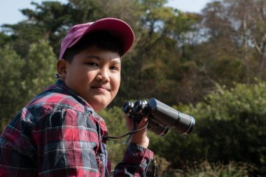 Asian boy held binoculars in hands and doing the bird watching, fish watching and forest observing in local national park, asian children summer vacation concept.
