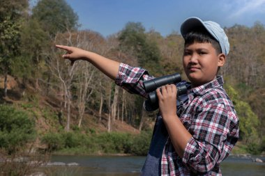 Asian boy held binoculars in hands and doing the bird watching, fish watching and forest observing in local national park, asian children summer vacation concept.
