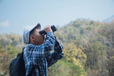 Asian boy held binoculars in hands and doing the bird watching, fish watching and forest observing in local national park, asian children summer vacation concept.