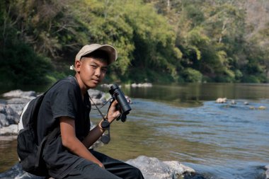 Asian boy held binoculars in hands and doing the bird watching, fish watching and forest observing in local national park, asian children summer vacation concept.