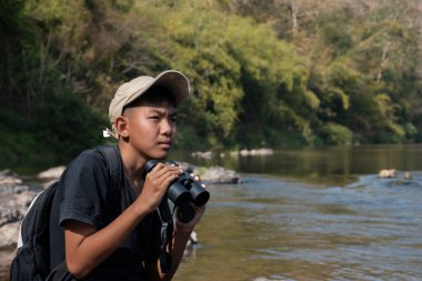 Asian boy held binoculars in hands and doing the bird watching, fish watching and forest observing in local national park, asian children summer vacation concept.