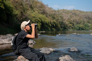 Asian boy held binoculars in hands and doing the bird watching, fish watching and forest observing in local national park, asian children summer vacation concept.