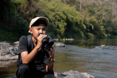 Asian boy held binoculars in hands and doing the bird watching, fish watching and forest observing in local national park, asian children summer vacation concept.