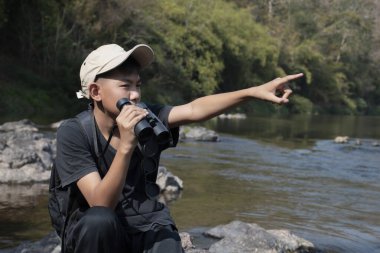 Asian boy held binoculars in hands and doing the bird watching, fish watching and forest observing in local national park, asian children summer vacation concept.