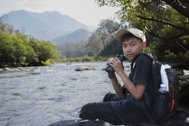 Asian boy held binoculars in hands and doing the bird watching, fish watching and forest observing in local national park, asian children summer vacation concept.
