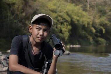 Asian boy held binoculars in hands and doing the bird watching, fish watching and forest observing in local national park, asian children summer vacation concept.