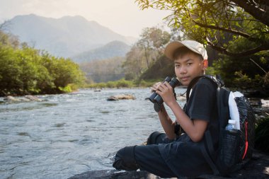 Asian boy held binoculars in hands and doing the bird watching, fish watching and forest observing in local national park, asian children summer vacation concept.
