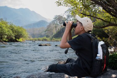Asian boy held binoculars in hands and doing the bird watching, fish watching and forest observing in local national park, asian children summer vacation concept.