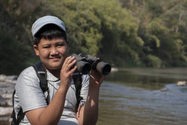 Asian boy held binoculars in hands and doing the bird watching, fish watching and forest observing in local national park, asian children summer vacation concept.