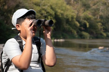 Asian boy held binoculars in hands and doing the bird watching, fish watching and forest observing in local national park, asian children summer vacation concept.