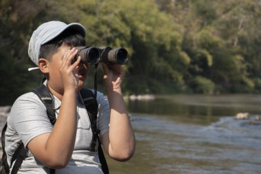 Asian boy held binoculars in hands and doing the bird watching, fish watching and forest observing in local national park, asian children summer vacation concept.