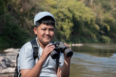 Asian boy held binoculars in hands and doing the bird watching, fish watching and forest observing in local national park, asian children summer vacation concept.
