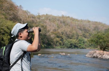 Asian boy held binoculars in hands and doing the bird watching, fish watching and forest observing in local national park, asian children summer vacation concept.