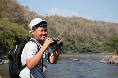 Asian boy held binoculars in hands and doing the bird watching, fish watching and forest observing in local national park, asian children summer vacation concept.