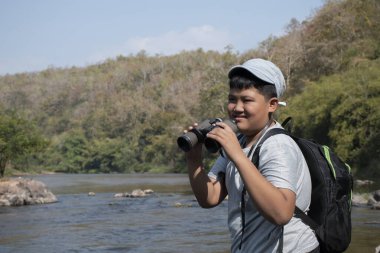 Asian boy held binoculars in hands and doing the bird watching, fish watching and forest observing in local national park, asian children summer vacation concept.