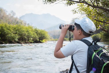 Asian boy held binoculars in hands and doing the bird watching, fish watching and forest observing in local national park, asian children summer vacation concept.