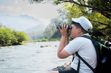 Asian boy held binoculars in hands and doing the bird watching, fish watching and forest observing in local national park, asian children summer vacation concept.