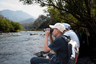 Asian boy held binoculars in hands and doing the bird watching, fish watching and forest observing in local national park, asian children summer vacation concept.