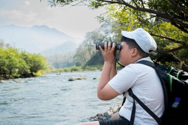 Asian boy held binoculars in hands and doing the bird watching, fish watching and forest observing in local national park, asian children summer vacation concept.
