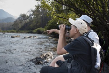 Asian boy held binoculars in hands and doing the bird watching, fish watching and forest observing in local national park, asian children summer vacation concept.