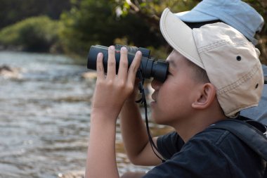 Asian boy held binoculars in hands and doing the bird watching, fish watching and forest observing in local national park, asian children summer vacation concept.
