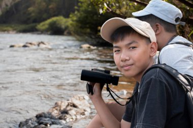 Asian boy held binoculars in hands and doing the bird watching, fish watching and forest observing in local national park, asian children summer vacation concept.