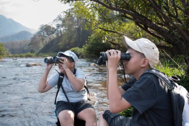 Asian boy held binoculars in hands and doing the bird watching, fish watching and forest observing in local national park, asian children summer vacation concept.
