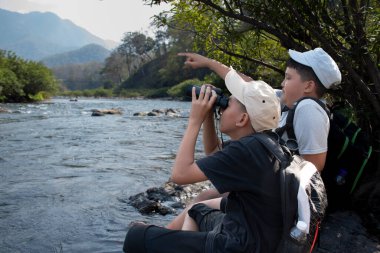 Asian boy held binoculars in hands and doing the bird watching, fish watching and forest observing in local national park, asian children summer vacation concept.