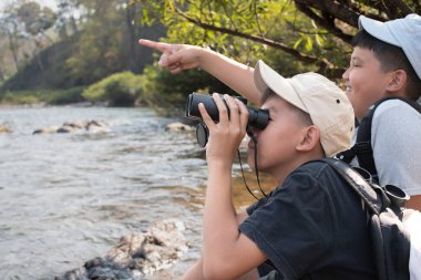 Asian boy held binoculars in hands and doing the bird watching, fish watching and forest observing in local national park, asian children summer vacation concept.