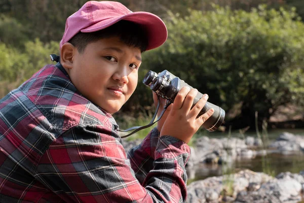 Asian boy held binoculars in hands and doing the bird watching, fish watching and forest observing in local national park, asian children summer vacation concept.