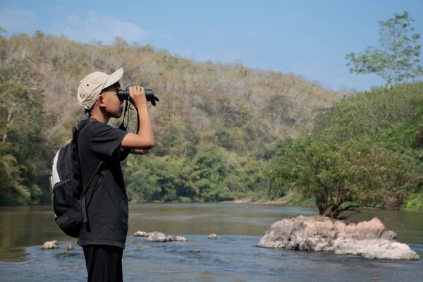 Asian boy held binoculars in hands and doing the bird watching, fish watching and forest observing in local national park, asian children summer vacation concept.