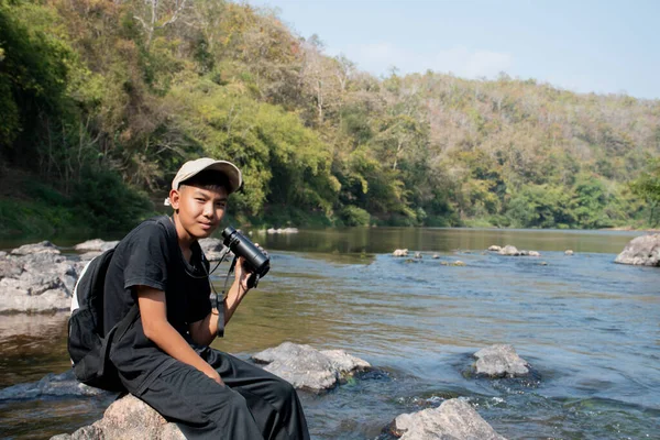 Asian boy held binoculars in hands and doing the bird watching, fish watching and forest observing in local national park, asian children summer vacation concept.