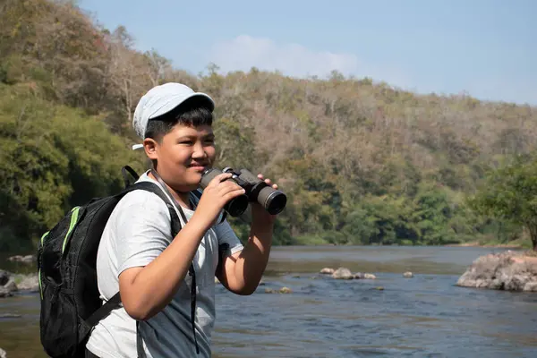 Asian boy held binoculars in hands and doing the bird watching, fish watching and forest observing in local national park, asian children summer vacation concept.