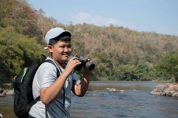 Asian boy held binoculars in hands and doing the bird watching, fish watching and forest observing in local national park, asian children summer vacation concept.