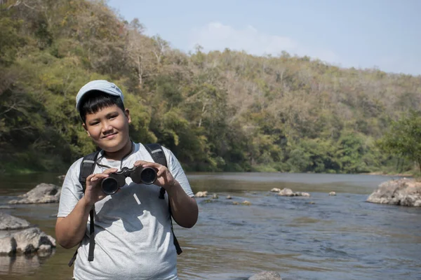 Asian boy held binoculars in hands and doing the bird watching, fish watching and forest observing in local national park, asian children summer vacation concept.
