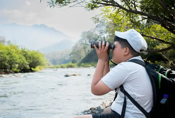 Asian boy held binoculars in hands and doing the bird watching, fish watching and forest observing in local national park, asian children summer vacation concept.
