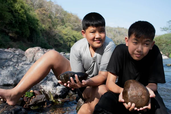 Asian boys're spending their freetimes by sitting on stones, holding rocks, scraping the top surface of stones to learn the natural life during their summer vacation in local national park.