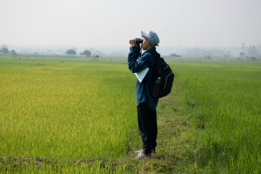 Asian boy in plaid shirt wears cap and has a backpack, holding and reading a map, standing on ridge rice field of asian farmers to observe the pm 2.5 smoke, dust and birds on tree branches and on sky.