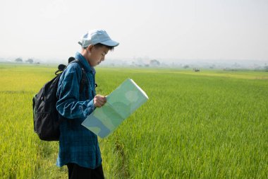 Asian boy in plaid shirt wears cap and has a backpack, holding and reading a map, standing on ridge rice field of asian farmers to observe the pm 2.5 smoke, dust and birds on tree branches and on sky.