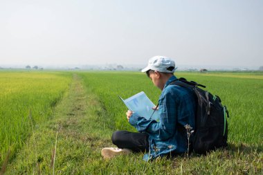 Asian boy wears plaid shirt and cap, has a backpack, binoculars and map, sitting on ridge of rice paddy field to observe the nature near village, birdwatching, pm 2.5 smoke, plants and rice growing.