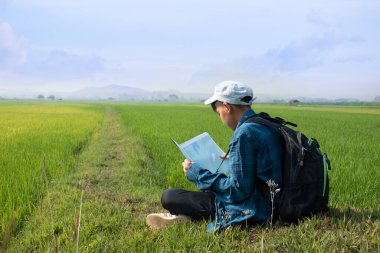 Asian boy wears plaid shirt and cap, has a backpack, binoculars and map, sitting on ridge of rice paddy field to observe the nature near village, birdwatching, pm 2.5 smoke, plants and rice growing.
