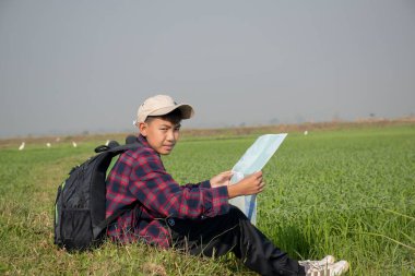 Asian boy in plaid shirt wears cap and has a backpack, holding a binoculars, sitting on ridge rice field of asian farmers to observe fish and birds on tree branches and on sky, soft focus.