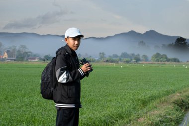 Asian boy wears cap and has a backpack, holding a binoculars, standing on ridge rice field of asian farmers to observe the pm 2.5 smoke, dust and birds on tree branches and on sky.