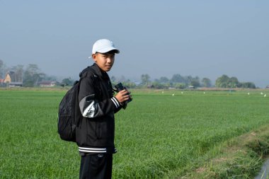 Asian boy wears cap and has a backpack, holding a binoculars, standing on ridge rice field of asian farmers to observe the pm 2.5 smoke, dust and birds on tree branches and on sky.