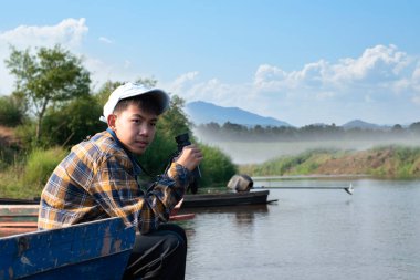 Asian boy in plaid shirt, holding a binoculars, sitting on boat which parked beside the local river to observe fish in the river and birds on tree branches and on sky.