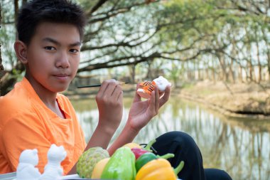 Asian boy in orange t-shirt sits on root of tree beside the pond in his  backyard garden and coloring tiger plaster doll during his weekend, recreational activity, relaxing and hobby at home concept.