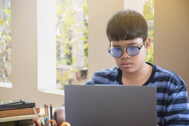 Asian boy wears eyesglasses, sitting near window in front of laptop and doing his online English exams seriously, concept for teenager daily lifestyle around the world.